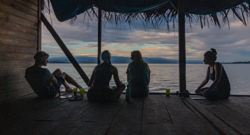 The silhouette of a group of people sitting on a dock at sunset appears before the ocean, with a body of land in the background. 